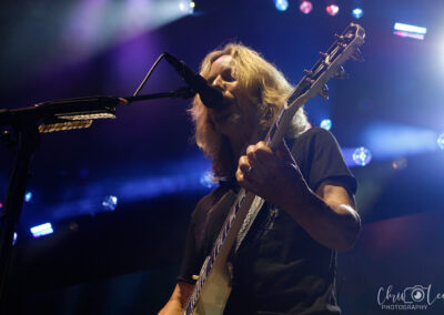 A musician with long hair plays an electric guitar and sings into a microphone on a brightly lit stage, with colorful lights and the logo Chris Lee Photography visible in the corner.