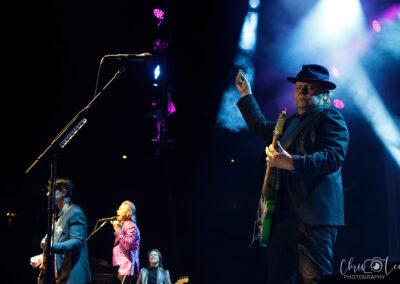 A band performs on stage under colorful lights; a guitarist in a hat raises his hand while other band members sing and play instruments in the background.