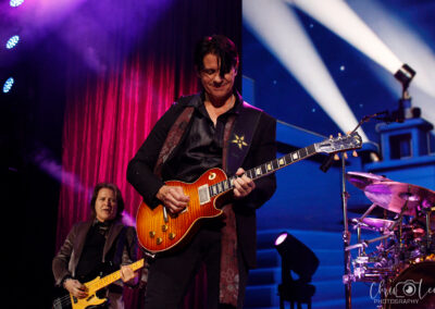 Two musicians perform on stage under purple lighting; the frontman plays an electric guitar while another plays a bass guitar behind him. Drums are visible on the right, with a backdrop that resembles a stylized night scene.