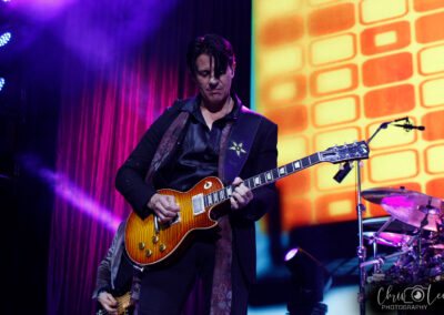 A man in a dark suit passionately plays an electric guitar on stage, with colorful lights and an orange geometric backdrop behind him. A drum set and another musician are partially visible.