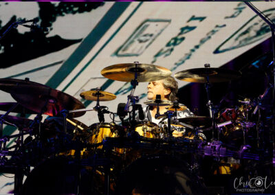 A drummer plays on a large drum kit under stage lights, with cymbals surrounding him and a colorful graphic displayed on the screen behind. The photo is credited to Chad Lee Photography.