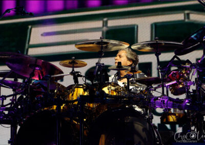 A drummer plays a large golden drum set on stage, surrounded by multiple cymbals, with a colorful, out-of-focus background and bright stage lighting.