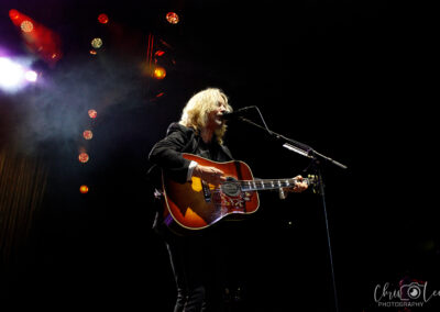 A musician with long blond hair plays an acoustic guitar and sings into a microphone on a dimly lit stage, with colorful lights and smoke in the background. The photo is credited to Chris Lee Photography.