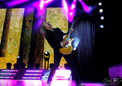 A guitarist with long hair passionately plays an electric guitar on stage, illuminated by purple and white lights, with a forest scene displayed on a screen behind him.