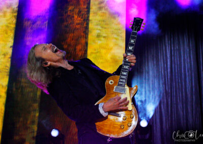 A guitarist with long hair passionately plays an electric guitar on stage, surrounded by colorful lights and smoke effects, creating a vibrant concert atmosphere.