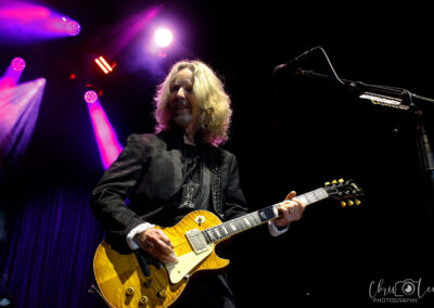 A musician with long blond hair plays an electric guitar on stage, illuminated by purple and pink stage lights, wearing a black jacket. A microphone and stand are visible on the right.