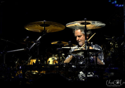 A drummer intensely plays a drum set on stage under dramatic lighting, surrounded by cymbals and drums, with shadows highlighting his concentration.