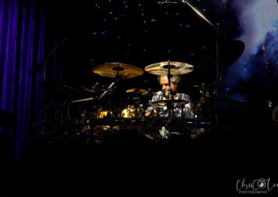 A drummer wearing sunglasses plays a drum kit on a dark stage, illuminated by spotlights, with a starry background behind him. The scene has a dramatic and energetic atmosphere.