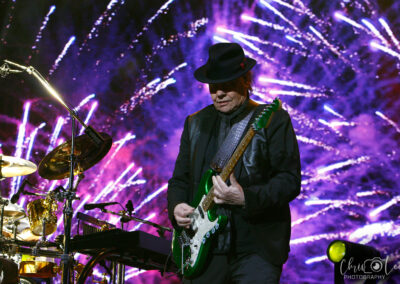 A musician in a black hat and suit plays a green electric guitar on stage, with vibrant purple and white fireworks displayed on a large screen in the background. Drums and stage equipment are visible beside him.