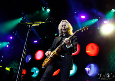 A guitarist with long blond hair plays an electric guitar on a stage with colorful, circular lights in the background. The scene is dramatic, with vibrant lighting and a professional concert atmosphere.