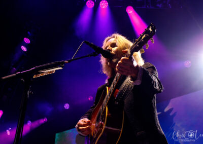 A musician with long blonde hair plays guitar and sings into a microphone on stage, illuminated by purple and pink concert lights.
