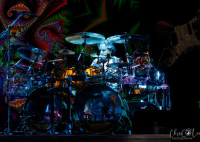 A drummer plays an elaborate drum kit on stage, surrounded by cymbals and drums, with a colorful psychedelic background projected behind them. The scene is vibrant and energetic, with dramatic lighting highlighting the performer.