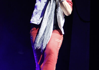 A person with long gray hair and sunglasses performs on stage, wearing a white vest, red pants, a gray scarf, and white shoes, holding a microphone and raising one arm under stage lights.