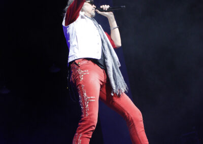 A singer wearing red pants, a white vest, and a scarf performs on stage, holding a microphone and raising one arm, with dramatic lighting and smoke in the background.