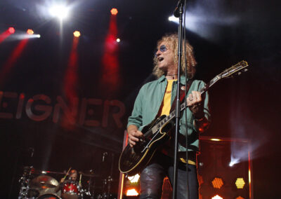 A guitarist with curly hair and sunglasses performs on stage with a black guitar, while a drummer plays in the background. The stage is lit with colorful lights, and the word Foreigner is partially visible behind them.