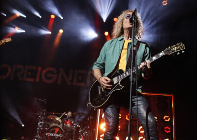 A musician with curly hair and sunglasses plays electric guitar and sings on stage under bright lights, with a drummer in the background and Foreigner visible on the drum kit and stage backdrop.