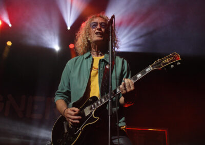 A musician with curly blond hair and glasses plays an electric guitar and sings into a microphone on stage, with dramatic stage lighting and beams of light in the background.