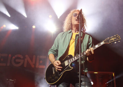 A musician with curly blond hair and sunglasses plays an electric guitar and sings into a microphone on a stage, with bright lights shining in the background.