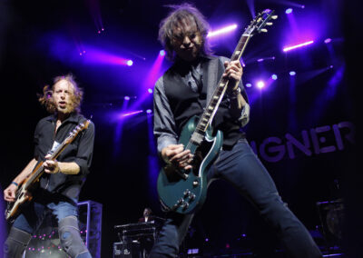 Two guitarists passionately perform on stage under vibrant purple lights, with the word FOREIGNER partially visible in the background. Both musicians are mid-action, intensively playing their electric guitars.