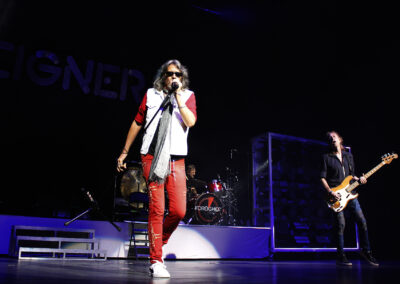 A rock band performs on stage with a lead singer in red pants and sunglasses at the center, a drummer in the background, and a bass guitarist to the right. The stage is dark with spotlights highlighting the musicians.
