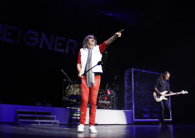 A rock band performs onstage under bright lights. The lead singer, wearing red pants and sunglasses, stands at the front, pointing toward the audience. A bassist plays in the background with the band name Foreigner visible behind them.