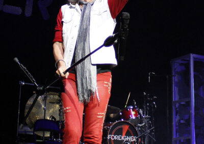 A rock singer wearing a white vest, red pants, and sunglasses performs on stage, holding a microphone stand and pointing forward. A drum set with Foreigner written on it is visible in the background.