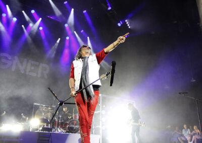 A rock singer in red pants and a denim vest performs energetically on stage, holding a microphone stand and pointing to the crowd. Purple stage lights shine above, with FOREIGNER visible in the background. Band members play behind him.