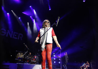 A rock band performs on stage under purple lights. The lead singer, wearing red pants and sunglasses, stands at the front with a microphone. A drummer and other band members play in the background. The word “FOREIGNER” is visible on stage.