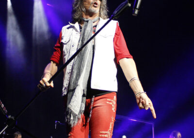 A male singer with long hair and sunglasses performs on stage, holding a microphone stand. He wears a white vest, red shirt, red pants, and a scarf. A drummer, band members, and Foreigner are visible in the background.