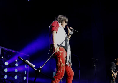 A singer in red pants and a white and red shirt performs passionately on stage with a microphone, dramatic blue and purple lighting, and a guitarist in the background.