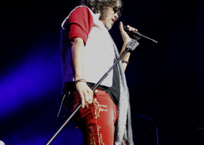 A singer in red pants, a white vest, and sunglasses holds a microphone on stage under dramatic blue and purple lighting.