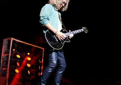 A man with curly hair wearing a teal shirt and dark pants plays an electric guitar on stage under bright lights, with a background of stage equipment and red lighting effects.