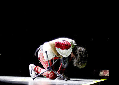 A performer with long hair, dressed in red pants and a white jacket, kneels on a stage, leaning forward over a microphone stand, illuminated by a spotlight against a dark background.