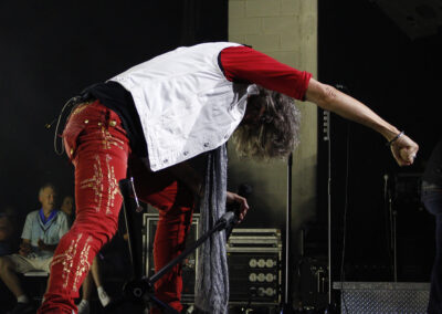 A performer in red pants and a white vest leans forward dramatically onstage, gripping a microphone stand, with audience members seated in the background.