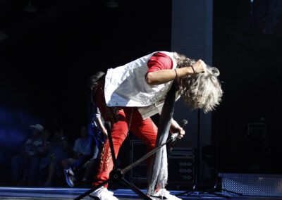 A performer with long, gray hair bends forward on stage, gripping a microphone stand. He wears red pants, a white vest, and a red shirt. Blue stage lights shine while audience members are visible in the background.