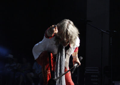 A performer with long gray hair kneels on stage under a spotlight, pointing toward the audience. They wear a white jacket, red pants, white sneakers, and a long gray scarf, holding a microphone in their left hand.