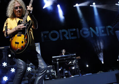 A guitarist with curly hair plays an electric guitar on stage under spotlights, with FOREIGNER illuminated in the background and a keyboard player performing behind him.
