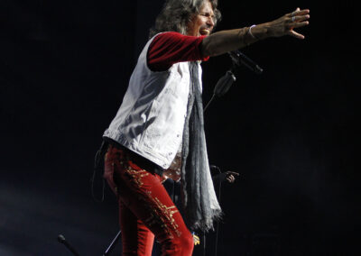 A male rock singer with long hair wears a white vest, red pants, and a gray scarf, performing passionately on stage with one arm extended toward the audience under dramatic lighting.