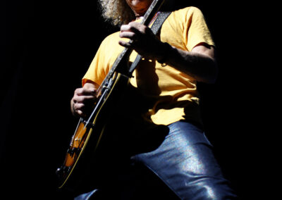 A musician with curly blond hair and sunglasses plays an electric guitar on stage, wearing a yellow shirt and blue jeans, dramatically lit against a dark background.
