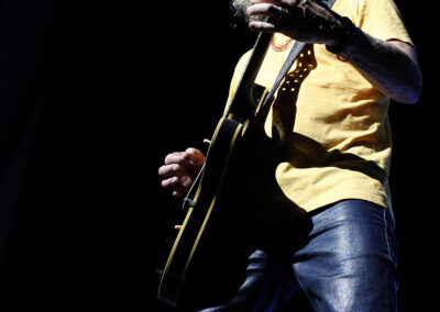 A musician with curly gray hair, wearing a yellow shirt and jeans, plays an electric guitar on a dark stage with dramatic lighting highlighting his face and instrument.