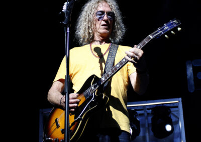 A man with long curly gray hair, wearing sunglasses and a yellow t-shirt, plays an electric guitar on stage under bright concert lights.