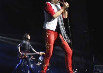 A rock singer in red pants and a white vest performs on stage with a microphone, while a guitarist plays in the background under dramatic stage lighting.