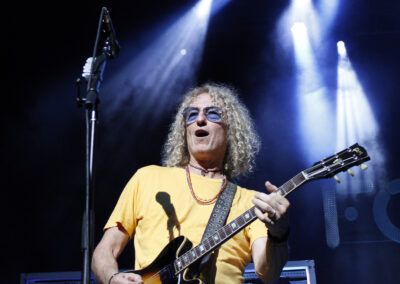 A musician with curly hair wearing sunglasses and a yellow shirt plays an electric guitar on stage under bright blue and white stage lights.