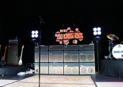 A stage setup for a concert with VOX amplifiers, a bass guitar, a drum set, stage lights, and a sign reading The Boxmasters in retro-style lettering.