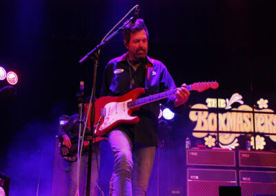 A musician plays an electric guitar on stage under purple lighting, with The Baymasters sign glowing in the background and guitar amplifiers stacked nearby.