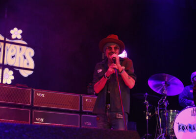 A singer wearing sunglasses and a hat performs on stage with a microphone, backed by amplifiers and a drummer with a drum set labeled The Boxmasters. Purple stage lighting and a retro sign are visible in the background.
