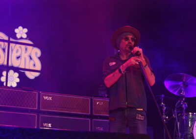 A man wearing sunglasses and a hat sings into a microphone on stage, surrounded by purple lighting, guitar amplifiers, and part of a drum set. A retro-style sign with the word SISTERS is visible in the background.