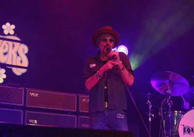 A singer wearing sunglasses and a hat performs on stage with a microphone. Amplifiers and a drum set are visible, and colorful lighting creates a vibrant concert atmosphere. The band’s name appears in retro-style lettering in the background.