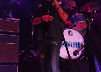 A musician wearing a hat, sunglasses, and casual clothes sings into a microphone on stage, with a drummer and drum set visible in the background under purple stage lights.