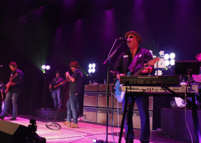 A band performs on stage under purple lights; one musician plays a keyboard and sings into a microphone, while others play guitars and drums. Large amplifiers and music equipment are visible in the background.